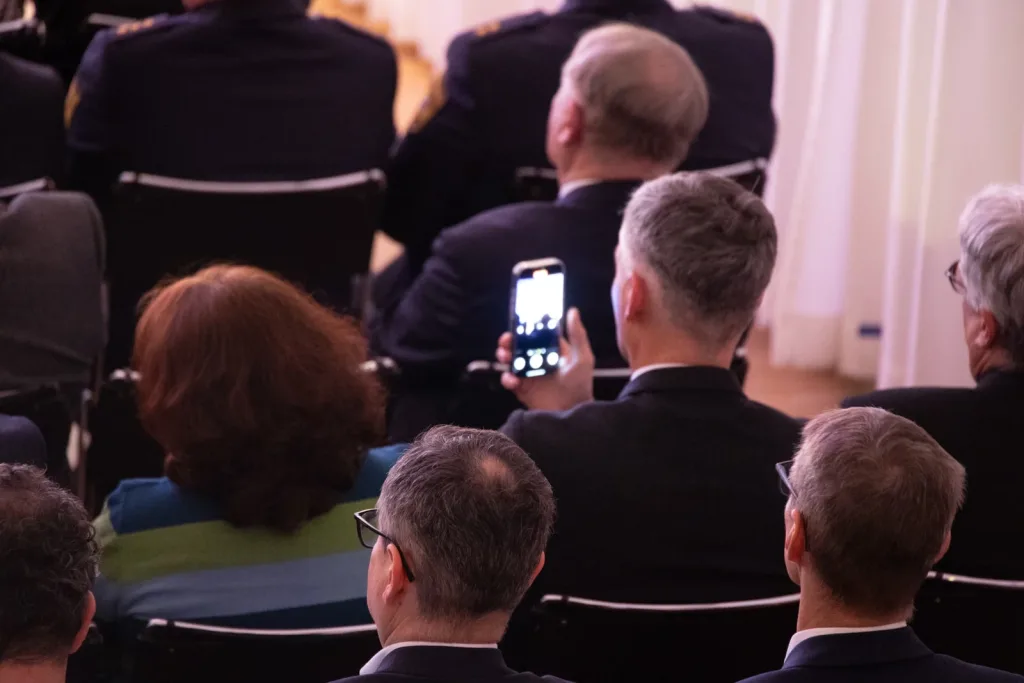 Gäste sitzen bei einem Neujahrsempfang in Reihen und fotografieren das Geschehen mit einem Smartphone.