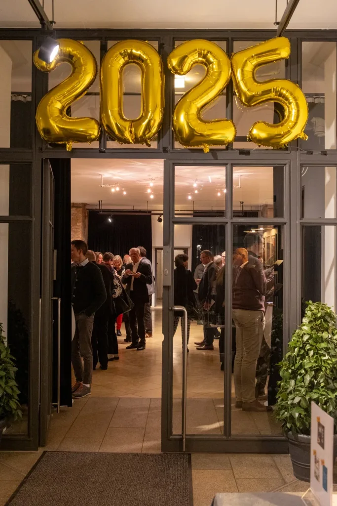 Eingangsbereich eines Empfangs mit großen goldenen Ballonzahlen „2025“ über den Glastüren und mehreren wartenden oder stehenden Gästen im Foyer
