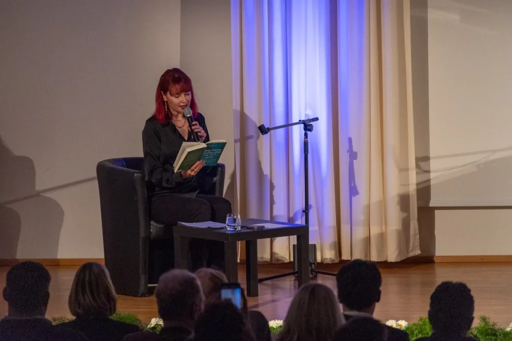 Eine Frau sitzt auf der Bühne, spricht in ein Mikrofon und hält ein Buch oder eine Mappe. Im Vordergrund sitzt Publikum.