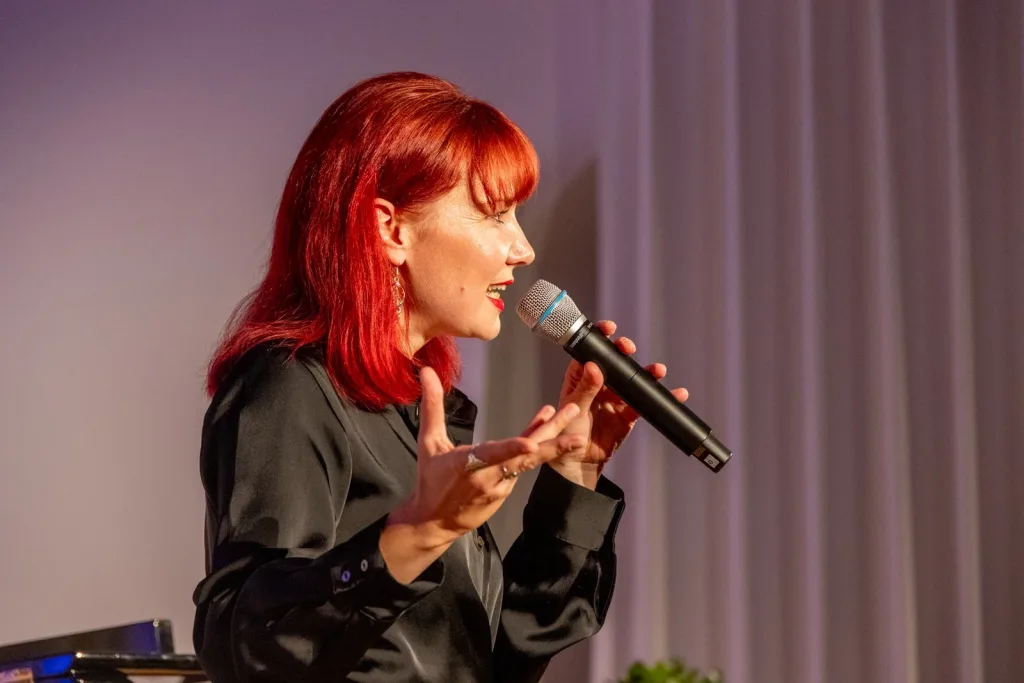 Seitlich aufgenommene Rednerin mit roten Haaren und schwarzer Kleidung spricht in ein Mikrofon.