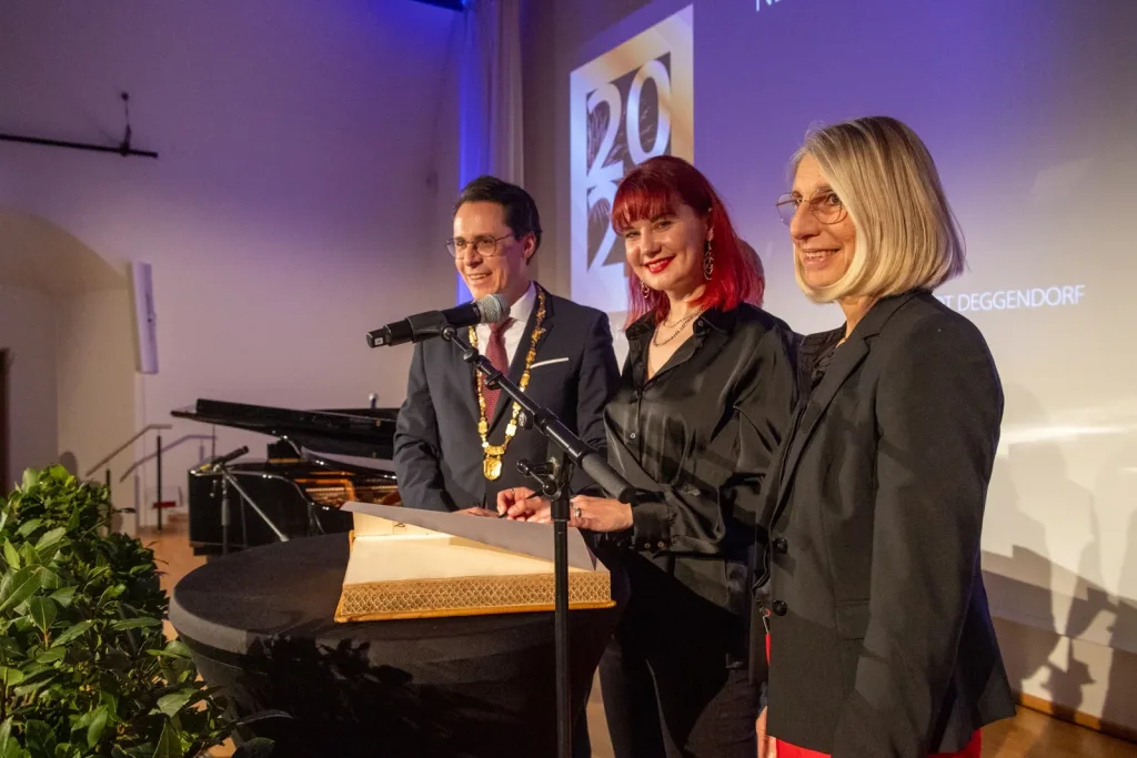 Drei Personen stehen bei einem Neujahrsempfang an einem Rednerpult neben einer angeschnittenen Torte; im Hintergrund sind eine Projektionsfläche und ein Flügel zu sehen.