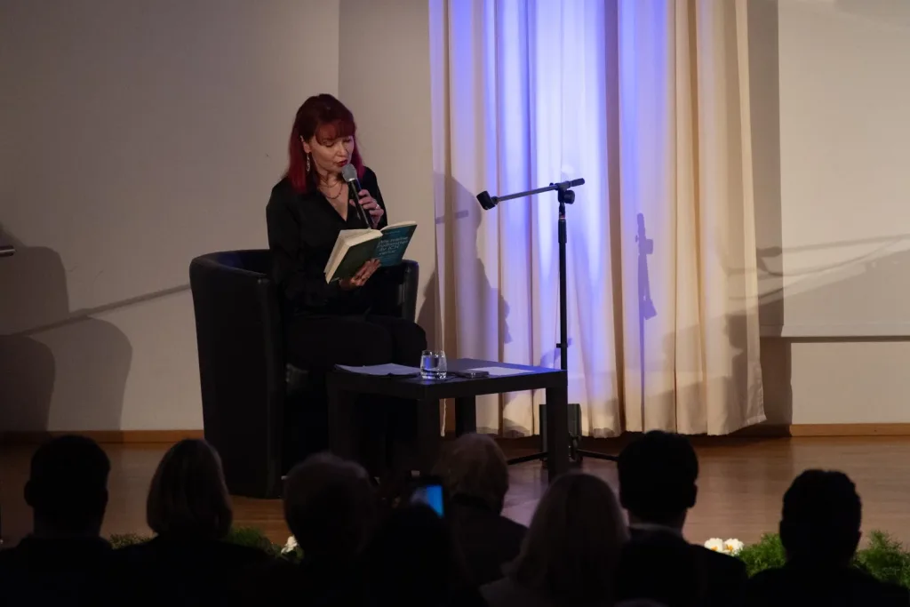 Frau liest auf einer Bühne aus einem Buch, während das Publikum im Vordergrund sitzt.