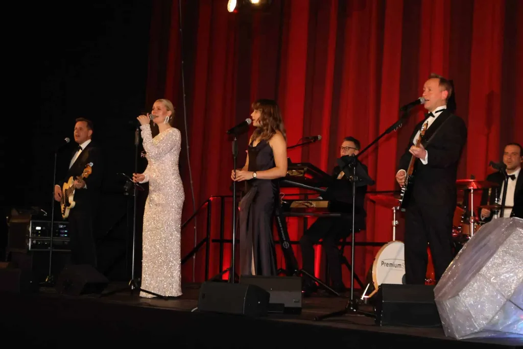 Vier Musikerinnen und Musiker spielen mit einer Sängerin und einem Sänger vor roten Vorhängen auf einer Bühne.