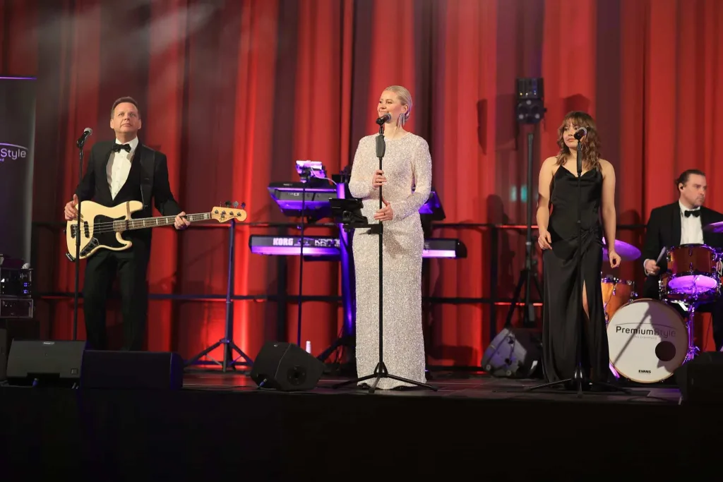 Vier Musikerinnen und Musiker treten vor roten Vorhängen auf einer Bühne auf; eine Sängerin steht am Mikrofon, daneben musizieren Gitarrist, Sängerin und Schlagzeuger.