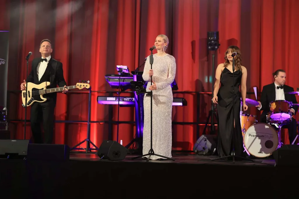 Eine Band mit zwei Sängerinnen und einem Sänger tritt vor roten Vorhängen auf einer beleuchteten Bühne auf.