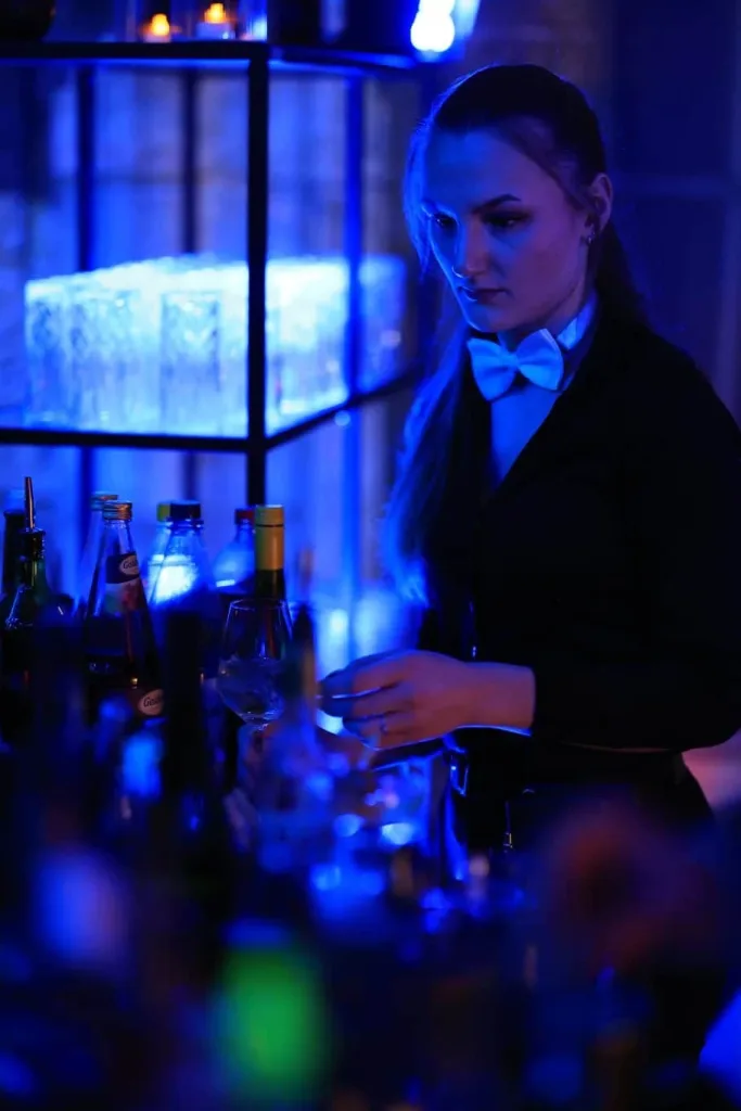 Barkeeperin im schwarzen Hemd mit Fliege bereitet hinter einer blau beleuchteten Bar ein Getränk zu.