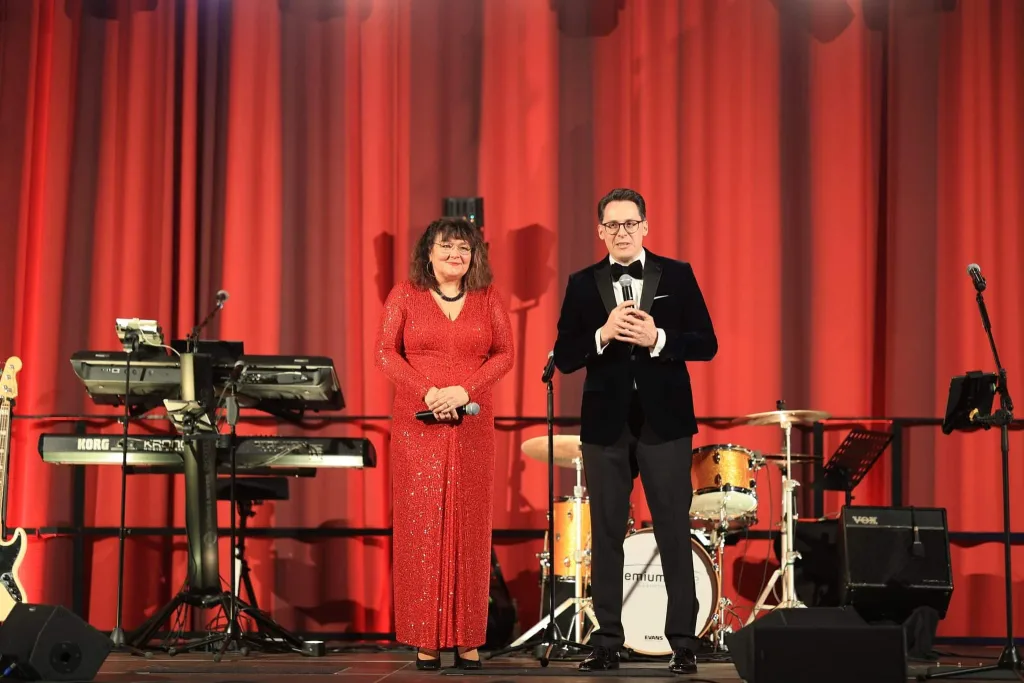 Zwei Personen stehen vor rotem Bühnenvorhang und Musikinstrumenten; der Mann hält ein Mikrofon, die Frau trägt ein rotes Kleid.