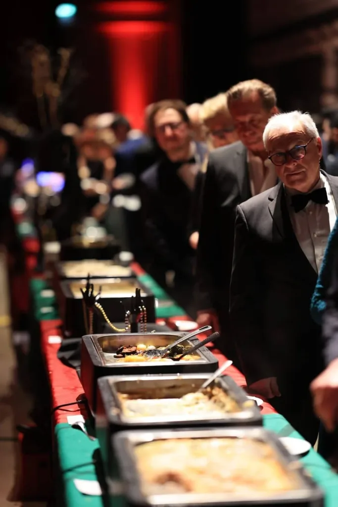 Gäste stehen an einem Buffet; im Vordergrund ist ein Mann im schwarzen Smoking mit Fliege zu sehen.