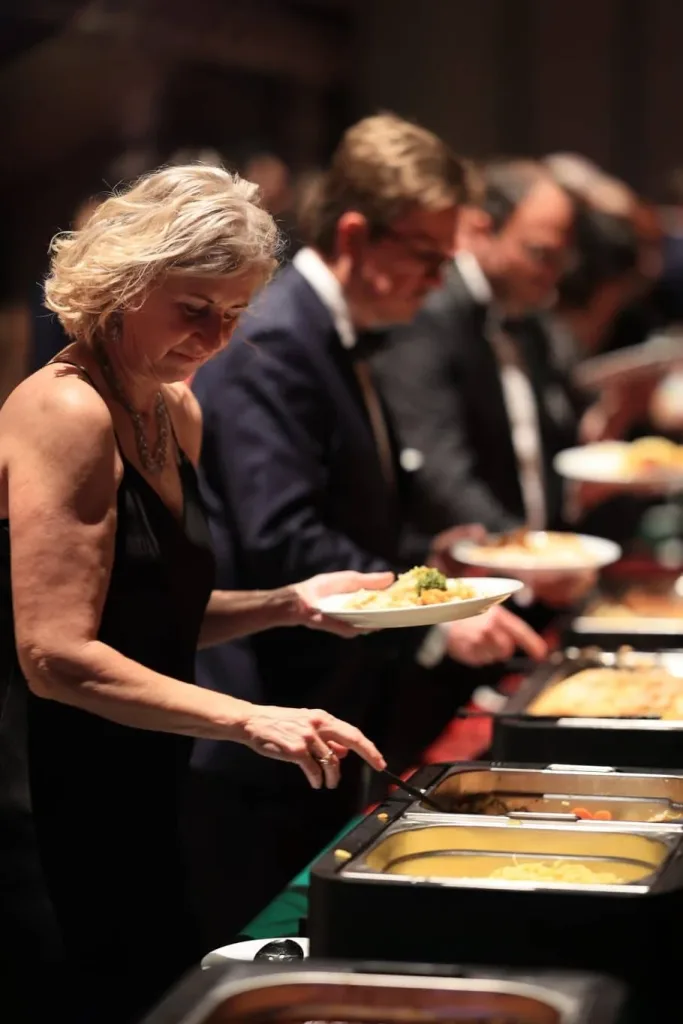 Gäste bedienen sich an einem Buffet; im Vordergrund hält eine Frau im schwarzen Kleid einen Teller und schöpft Speisen.