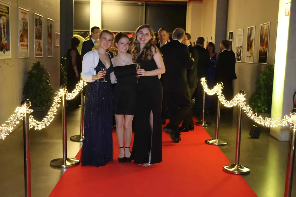 Drei festlich gekleidete junge Frauen posieren auf einem roten Teppich, während weitere Gäste durch einen beleuchteten Eingangsbereich gehen.