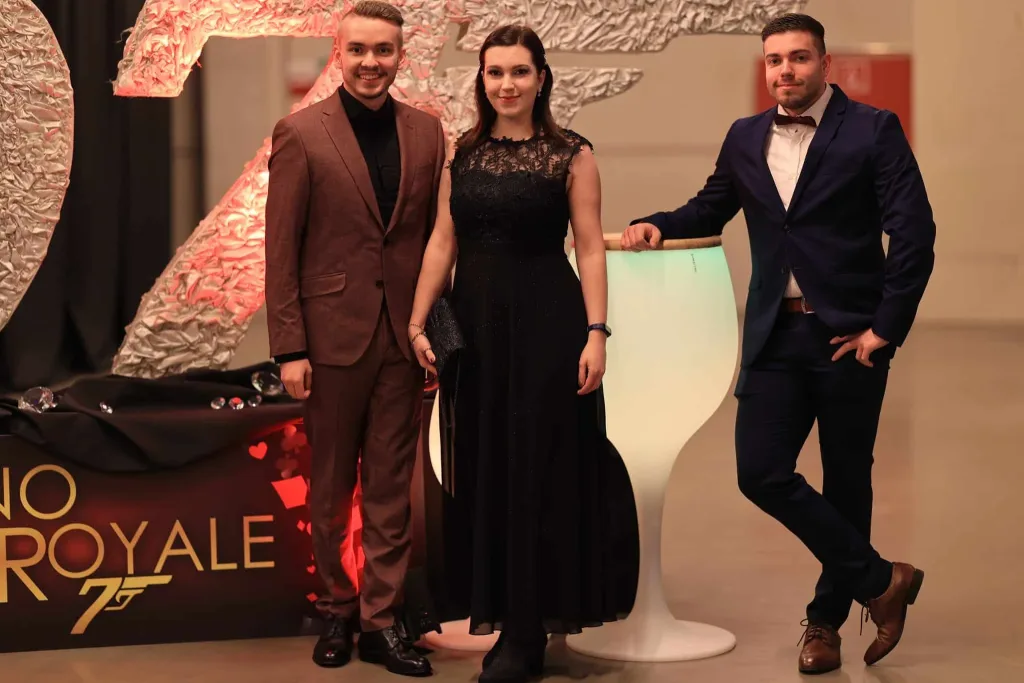 Drei elegant gekleidete Personen stehen neben einer beleuchteten Bar mit der Aufschrift „Casino Royale“.