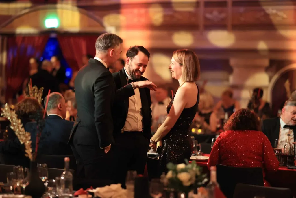 Drei elegant gekleidete Gäste stehen und unterhalten sich in einem festlich beleuchteten Saal, während weitere Personen an Tischen sitzen.