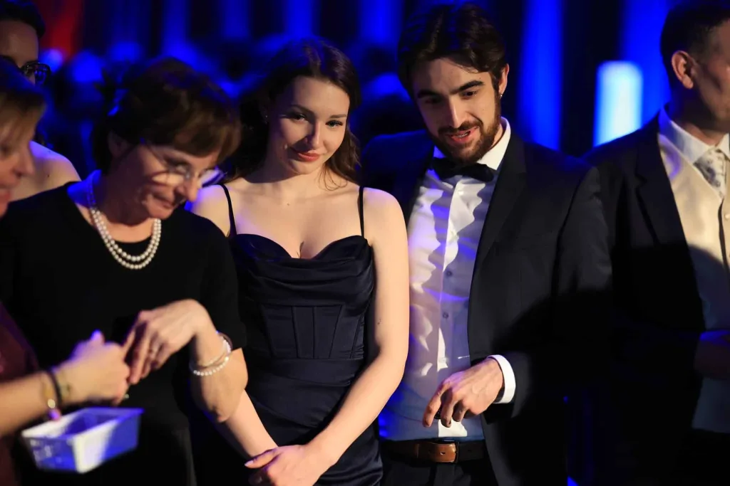 Mehrere elegant gekleidete Gäste stehen in blauem Licht beisammen; eine Frau trägt ein dunkles Kleid, ein Mann daneben einen schwarzen Smoking.