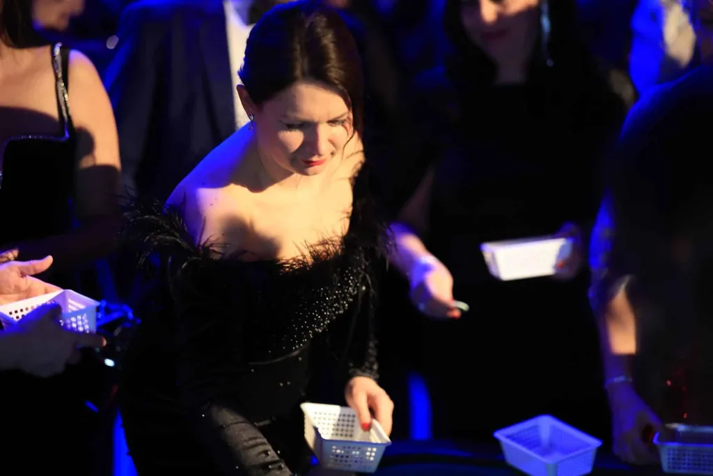Elegante Frau in schwarzer Kleidung hält ein kleines Körbchen an einem Tisch; im Hintergrund stehen weitere festlich gekleidete Menschen.