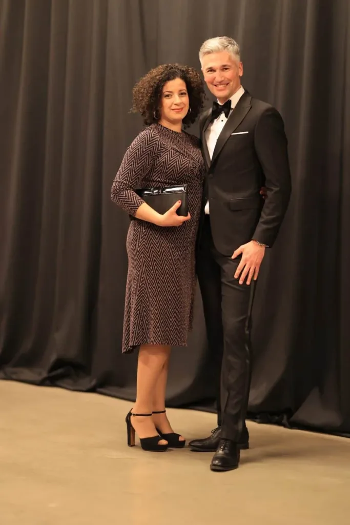 Eine Frau im gemusterten Kleid und ein Mann im schwarzen Smoking stehen vor einem dunklen Vorhang.