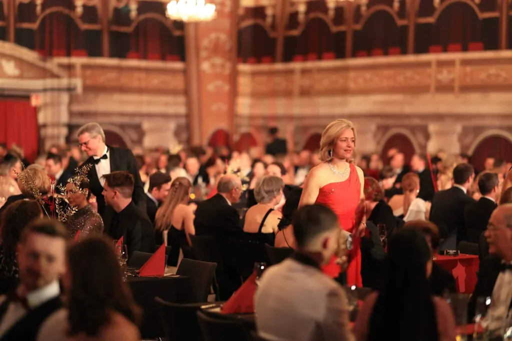 Festlich gekleidete Gäste sitzen und stehen in einem prunkvollen Saal, darunter eine Frau in einem roten Kleid.