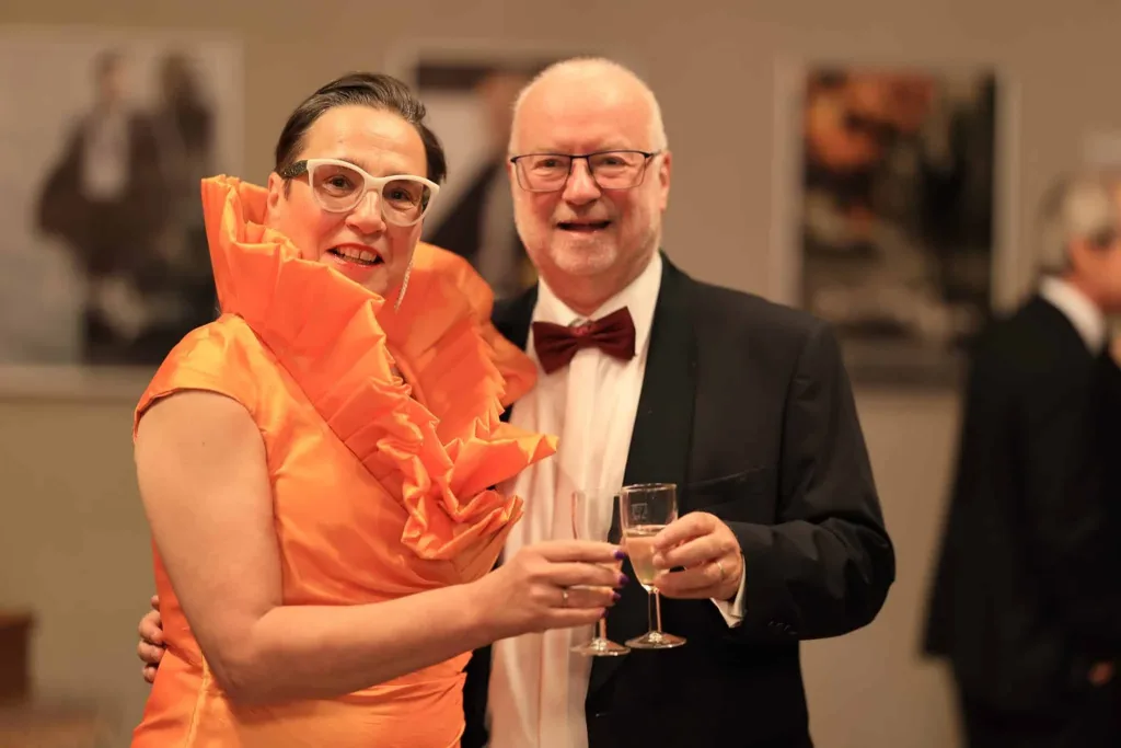 Eine Frau in einem leuchtend orangefarbenen Kleid und ein Mann im schwarzen Smoking stoßen mit Sektgläsern an.