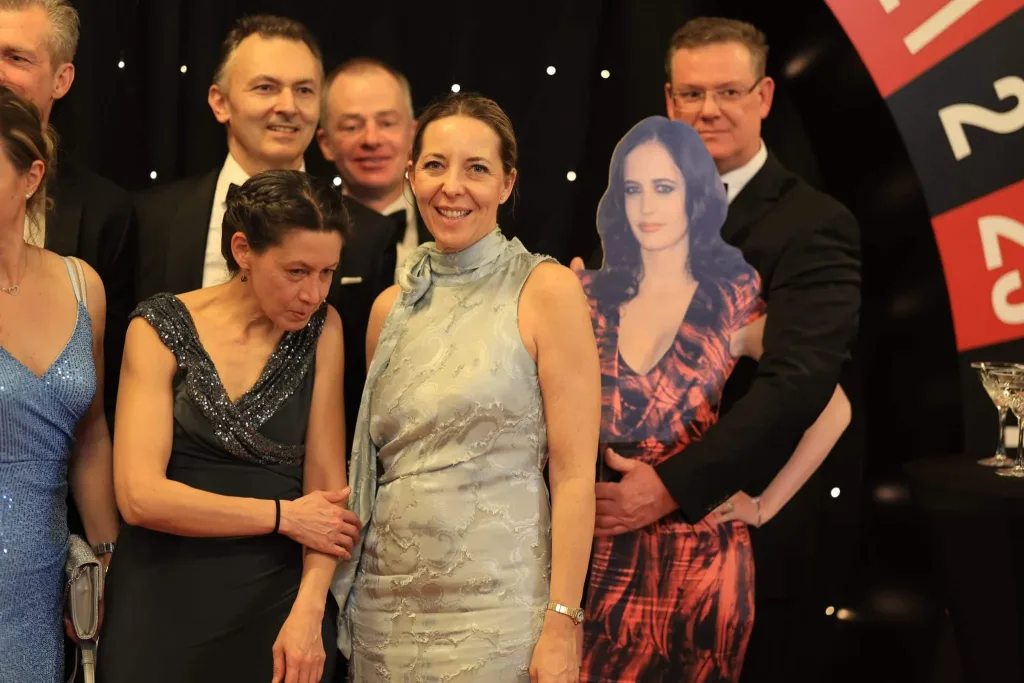 Mehrere festlich gekleidete Personen stehen vor einem schwarzen Hintergrund; rechts ist ein aufgestelltes Foto einer Frau zu sehen.