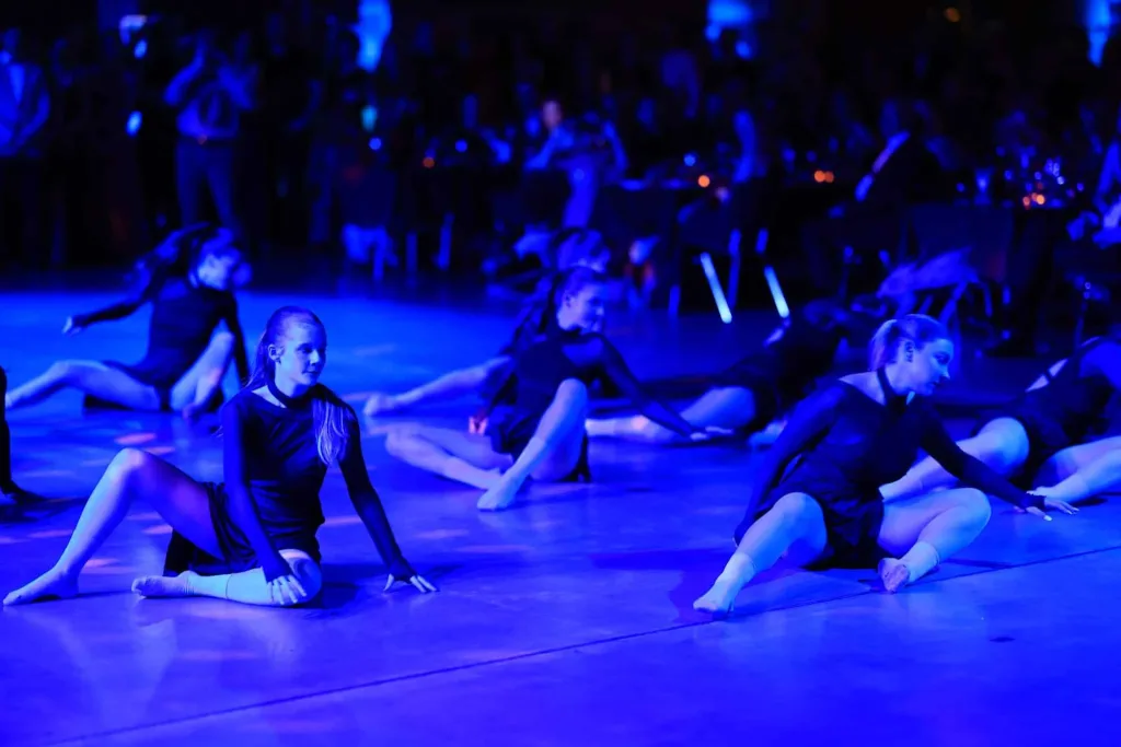 Mehrere Tänzerinnen in schwarzen Kostümen sitzen und bewegen sich bei blauem Licht synchron auf einer Tanzfläche.