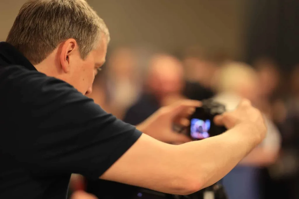 Fotograf mit grauem Haar fotografiert mit einer Kamera; im Hintergrund sind mehrere unscharfe Personen zu sehen.