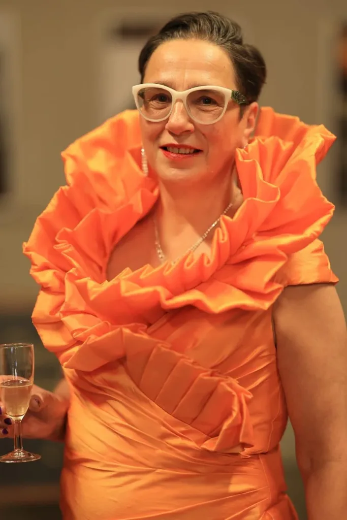 Frau mit Brille und auffälligem orangefarbenem Kleid hält ein Glas in der Hand.