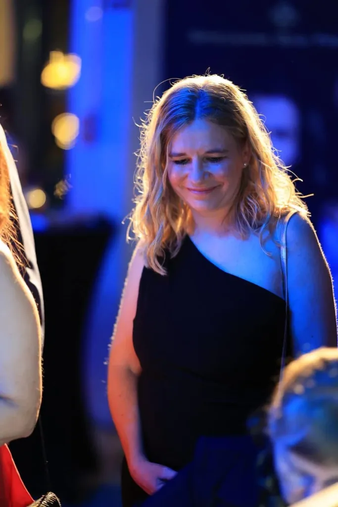 Eine blonde Frau in einem schwarzen Kleid steht bei blauer Beleuchtung in einem Innenraum.