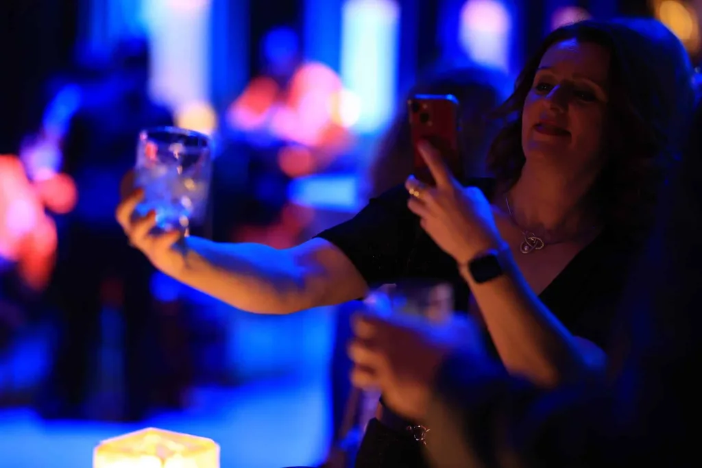 Frau hält ein Smartphone und ein Cocktailglas in einem blau beleuchteten Innenraum.