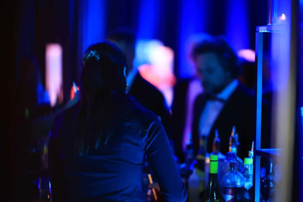 Unscharf beleuchtete Gäste stehen an einer Bar mit Getränkeflaschen und Gläsern.