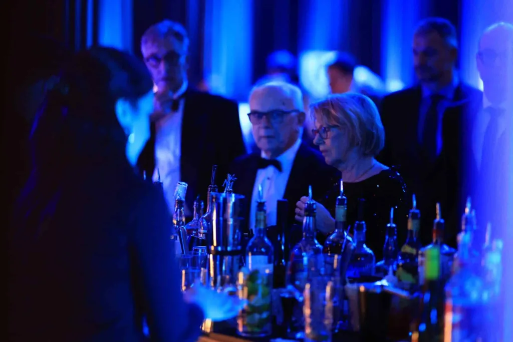 Mehrere Gäste sitzen und stehen an einer Bar mit Getränken in blauem Licht.