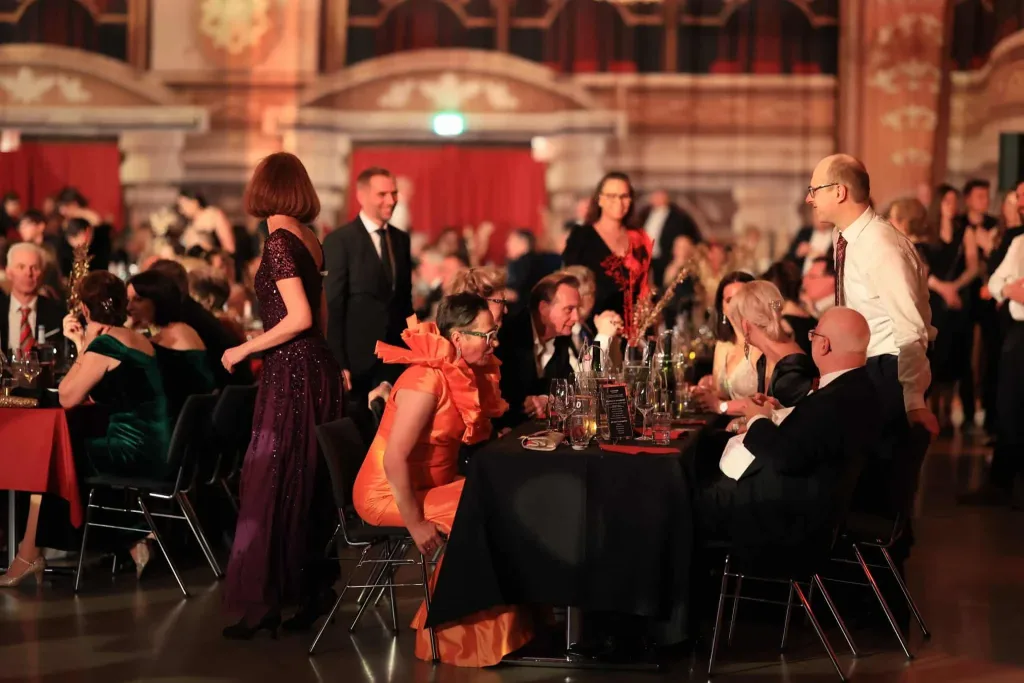 Gäste sitzen und stehen an festlich gedeckten Tischen in einem prunkvollen Saal; mehrere Personen tragen elegante Abendkleidung.