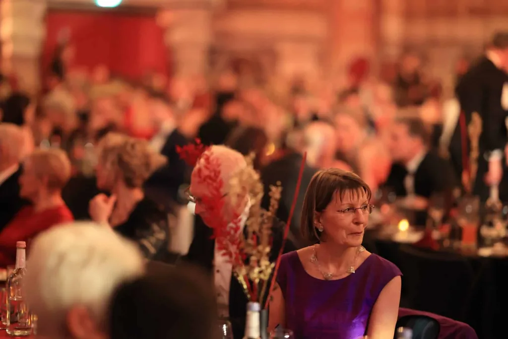 Frau im violetten Kleid sitzt an einem festlich gedeckten Tisch; im Hintergrund sitzen weitere Gäste in einem warm beleuchteten Saal.