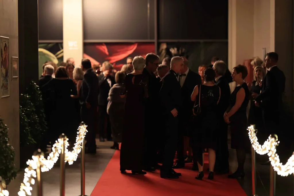 Elegant gekleidete Gäste stehen und gehen auf einem rot beleuchteten Teppich in einem festlich dekorierten Innenraum.