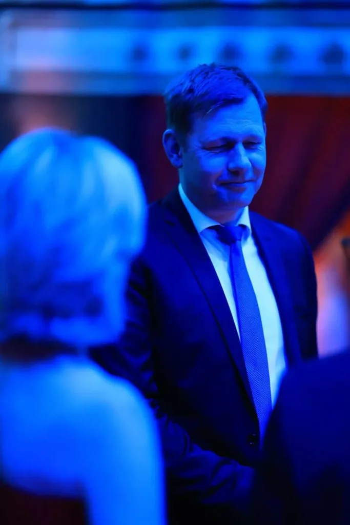 Eleganter Mann im dunklen Anzug steht bei blauem Licht; im Vordergrund ist unscharf der Rücken einer Person zu sehen.