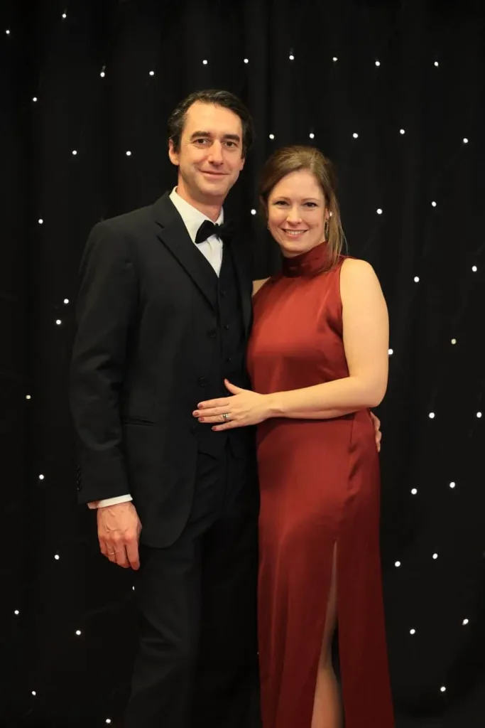 Ein Mann im schwarzen Anzug mit Fliege und eine Frau in einem roten Abendkleid stehen lächelnd vor einem schwarzen Hintergrund mit kleinen Lichtpunkten.