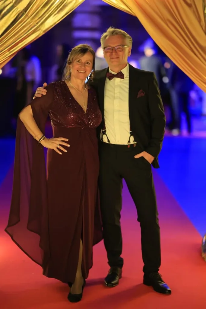 Eine Frau im dunkelroten Abendkleid und ein Mann im schwarzen Anzug mit Fliege stehen unter goldenen Vorhängen auf einem roten Teppich.