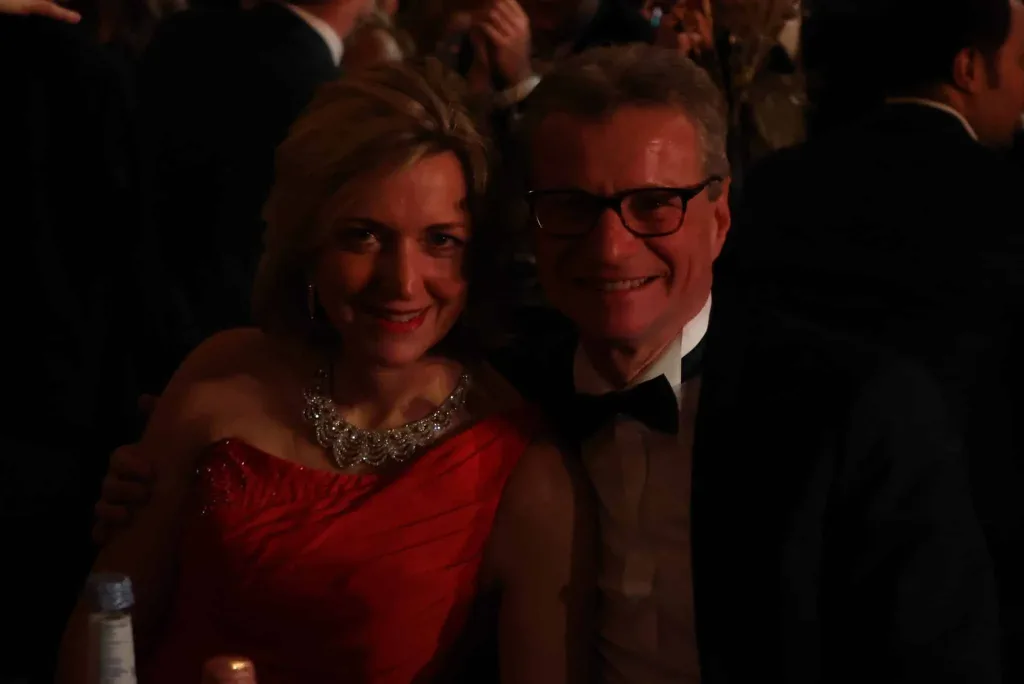 Eine lächelnde Frau in rotem Kleid und ein Mann mit Brille und schwarzer Fliege sitzen oder stehen dicht beieinander in einem abgedunkelten Raum.