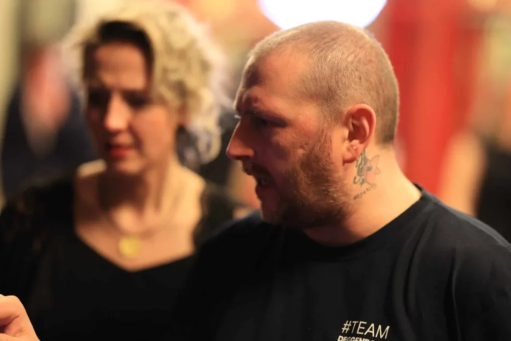 Seitliches Porträt eines bärtigen Mannes mit tätowiertem Hals und schwarzem T-Shirt; dahinter steht eine unscharfe Frau.