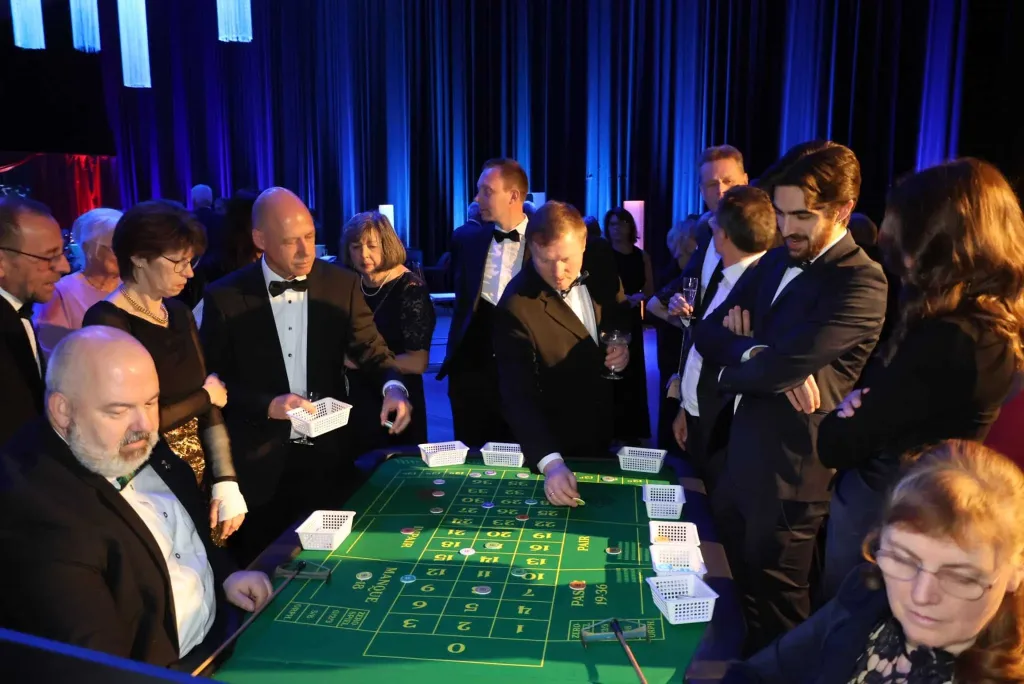 Gäste in festlicher Abendkleidung stehen und sitzen an einem Roulettetisch mit grüner Spielfläche und Jetons.