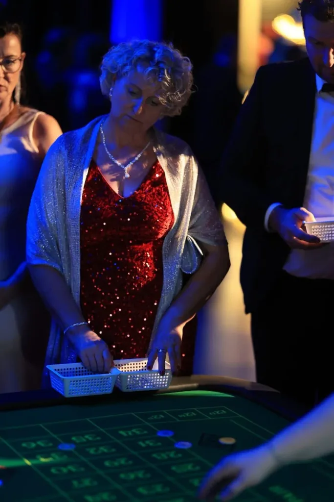 Ältere Frau in rotem Paillettenkleid und silberner Stola legt Jetons an einem Roulette-Tisch ab.