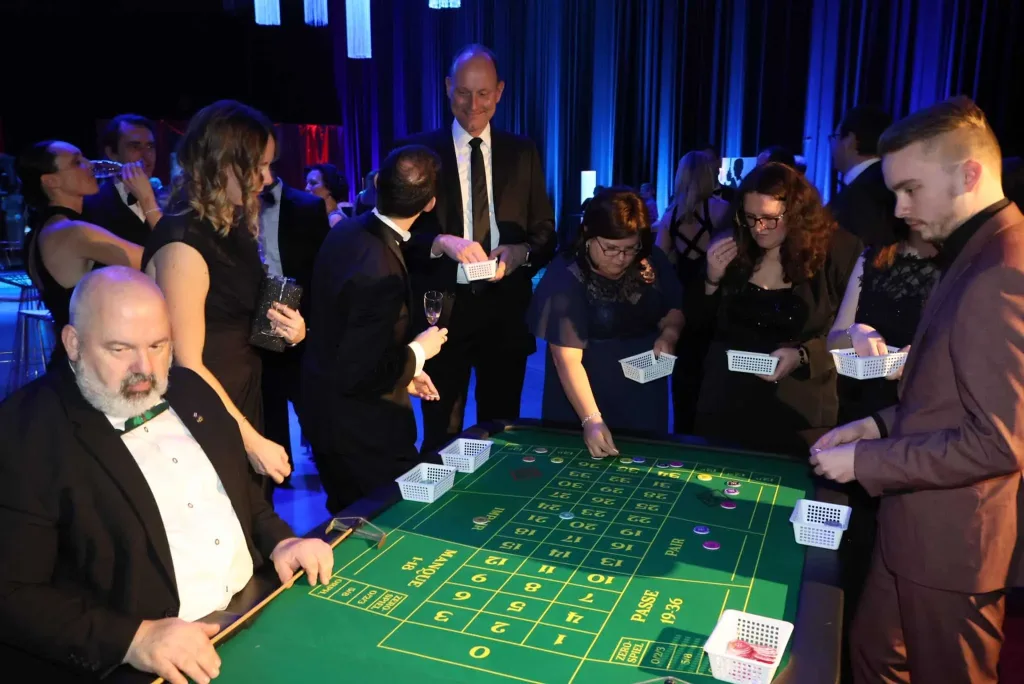 Gäste stehen und sitzen an einem Roulette-Tisch mit Spielkarten und Jetons in einem blau beleuchteten Saal.