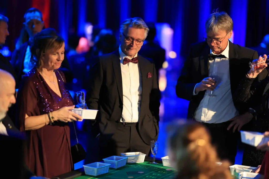 Mehrere festlich gekleidete Gäste stehen an einem Roulette- oder Spieltisch in blau beleuchteter Umgebung.