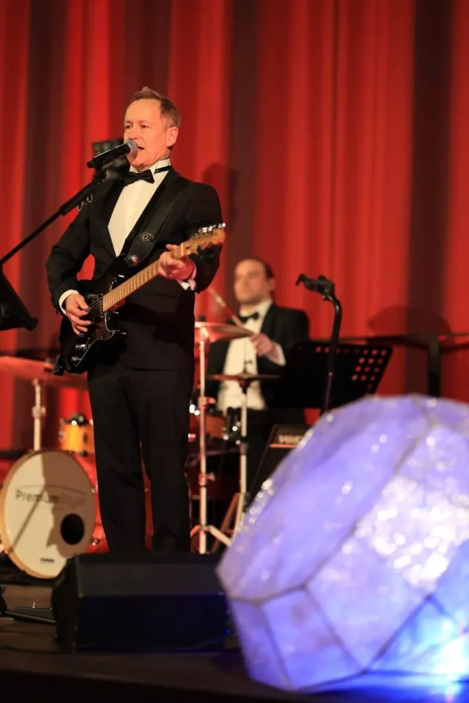 Sänger mit E-Gitarre singt vor roten Vorhängen, während ein Schlagzeuger im Hintergrund spielt.