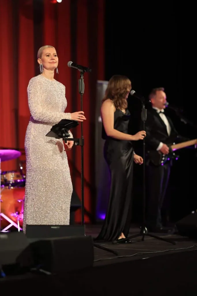 Sängerin in einem glitzernden langen Kleid steht an einem Mikrofon auf einer beleuchteten Bühne; dahinter musizieren zwei weitere Personen.