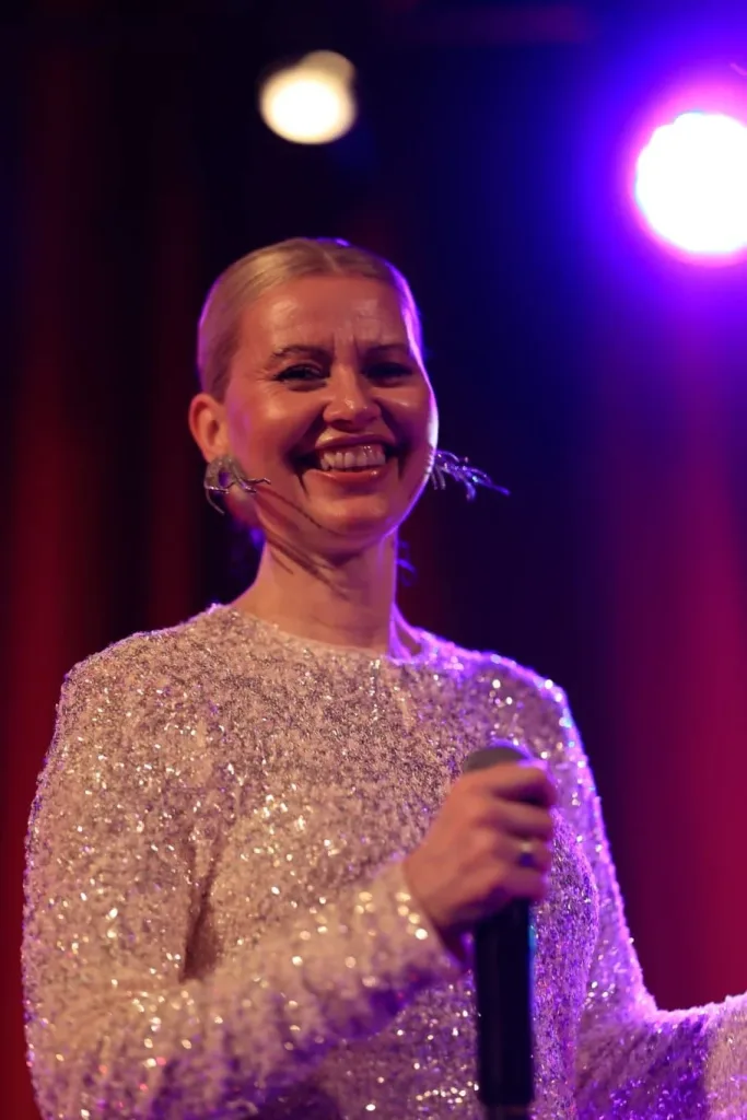 Lächelnde Sängerin mit Mikrofon und funkelndem Glitzerkleid auf einer farbig beleuchteten Bühne