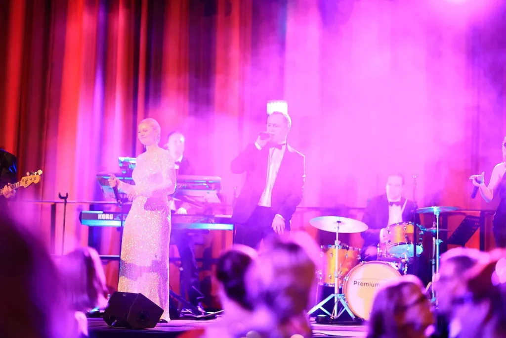Sängerin in einem glitzernden Kleid und Sänger mit Mikrofon auf einer farbig beleuchteten Bühne, begleitet von einer Band; davor Publikum
