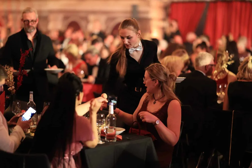 Eine Servicekraft schenkt an einem festlich gedeckten Tisch einer sitzenden Frau ein Getränk ein, während weitere Gäste im Hintergrund sitzen.