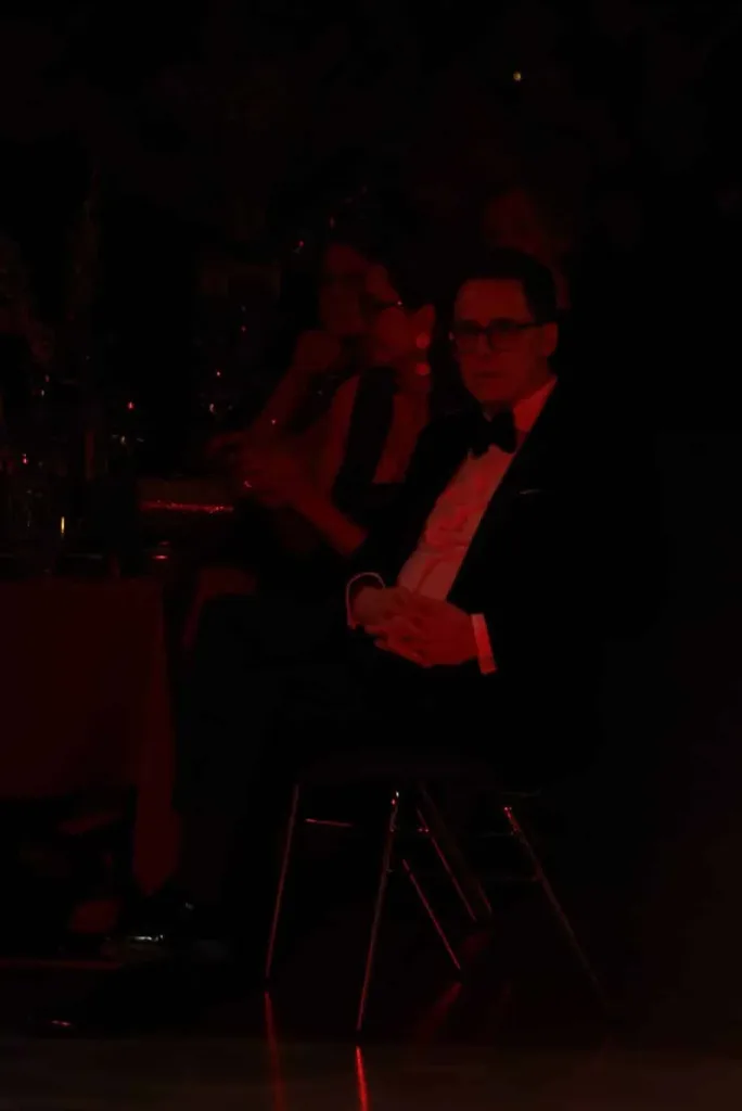 Sitzender Mann mit Brille im schwarzen Smoking und Fliege in einer dunkel beleuchteten Innenraumszene