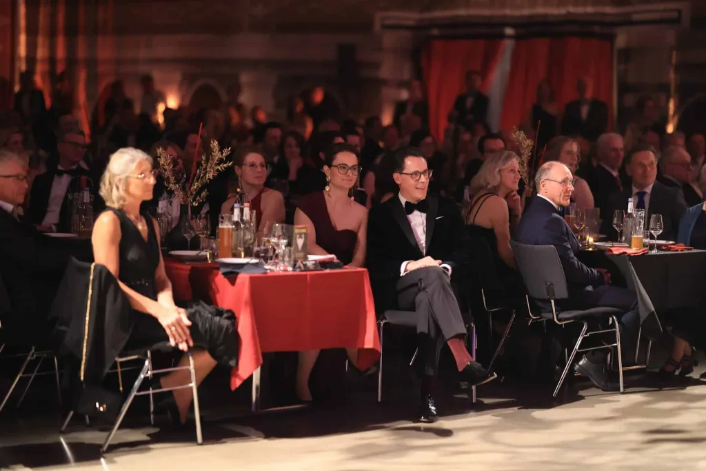 Elegantes Publikum sitzt an festlich gedeckten Tischen in einem abgedunkelten Saal.