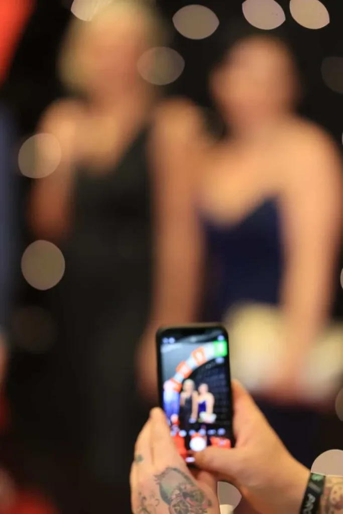 Eine Hand mit tätowiertem Unterarm hält ein Smartphone mit sichtbarer Kameraansicht; dahinter stehen zwei unscharfe Personen vor dunklem Hintergrund mit hellen Lichtkreisen.
