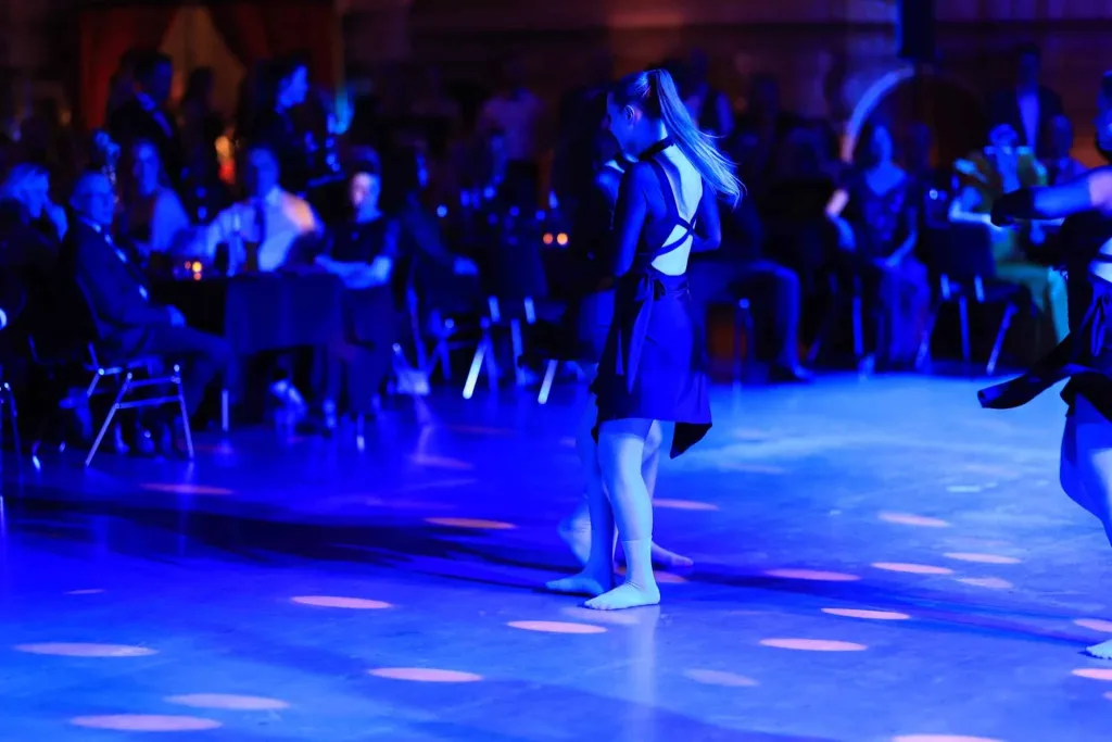 Tänzerin in dunklem Kleid steht auf einer blau beleuchteten Tanzfläche, während im Hintergrund Gäste sitzen oder stehen.